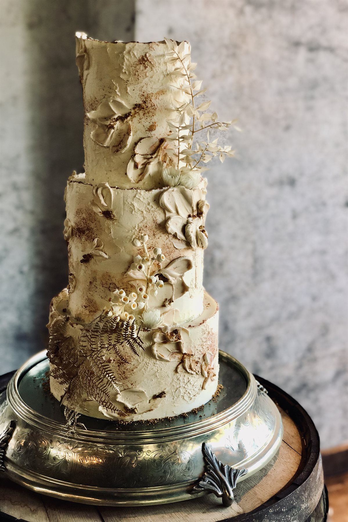 Three-tier textured buttercream cake with palette knife detail, dried flowers, gold-dusted fern and gypsophila on ornate silver barrel stand in a real venue