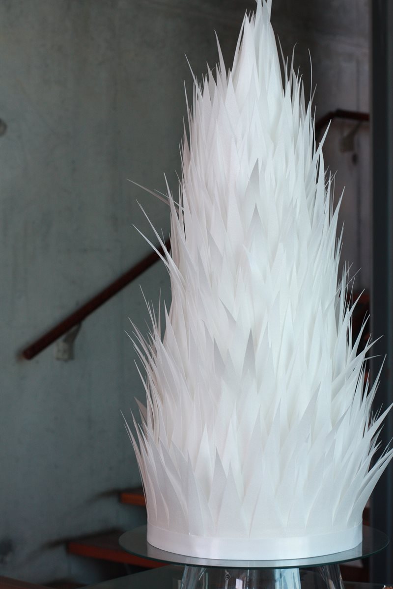 Tall multi-tier white contemporary cake covered entirely in countless hand-cut white feathered wafer paper spikes rising into a dramatic vertical spire, on a glass cake stand