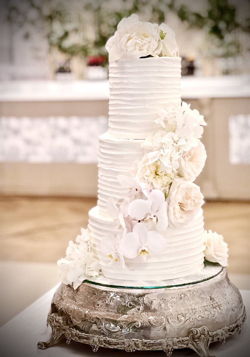 Three-tier horizontal-ribbed white buttercream cake with cascading white peonies and orchids in a real wedding venue