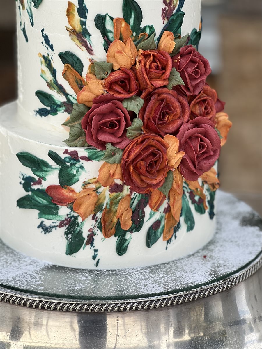 Close detail of a tiered cake hand-painted in autumnal forest-green, rust orange and burgundy brushwork, with sculpted buttercream roses and leaves in the same warm autumn tones, on a silver tray