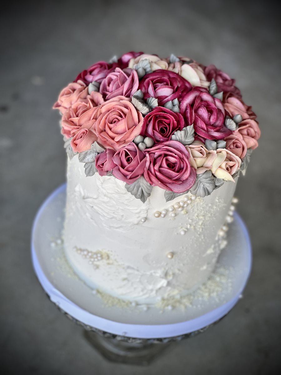 Tall single-tier white celebration cake topped with a bouquet of buttercream roses in pink, burgundy and cream, with delicate pearl accents at the base