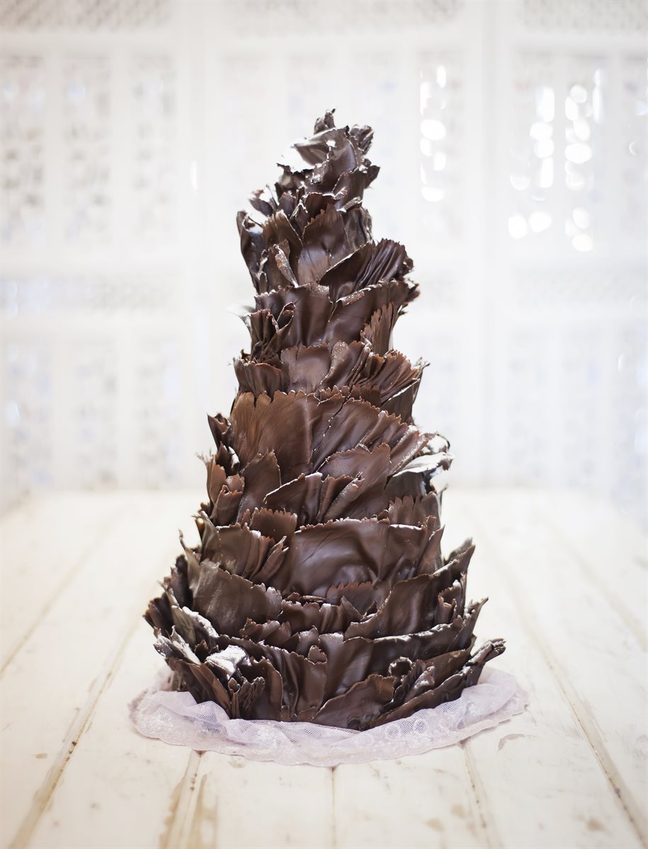 Tall dramatic dark couverture chocolate sculpted spire — layered hand-cut chocolate petals stacked into a towering cone, on a soft white wooden floor against a luminous lattice backdrop