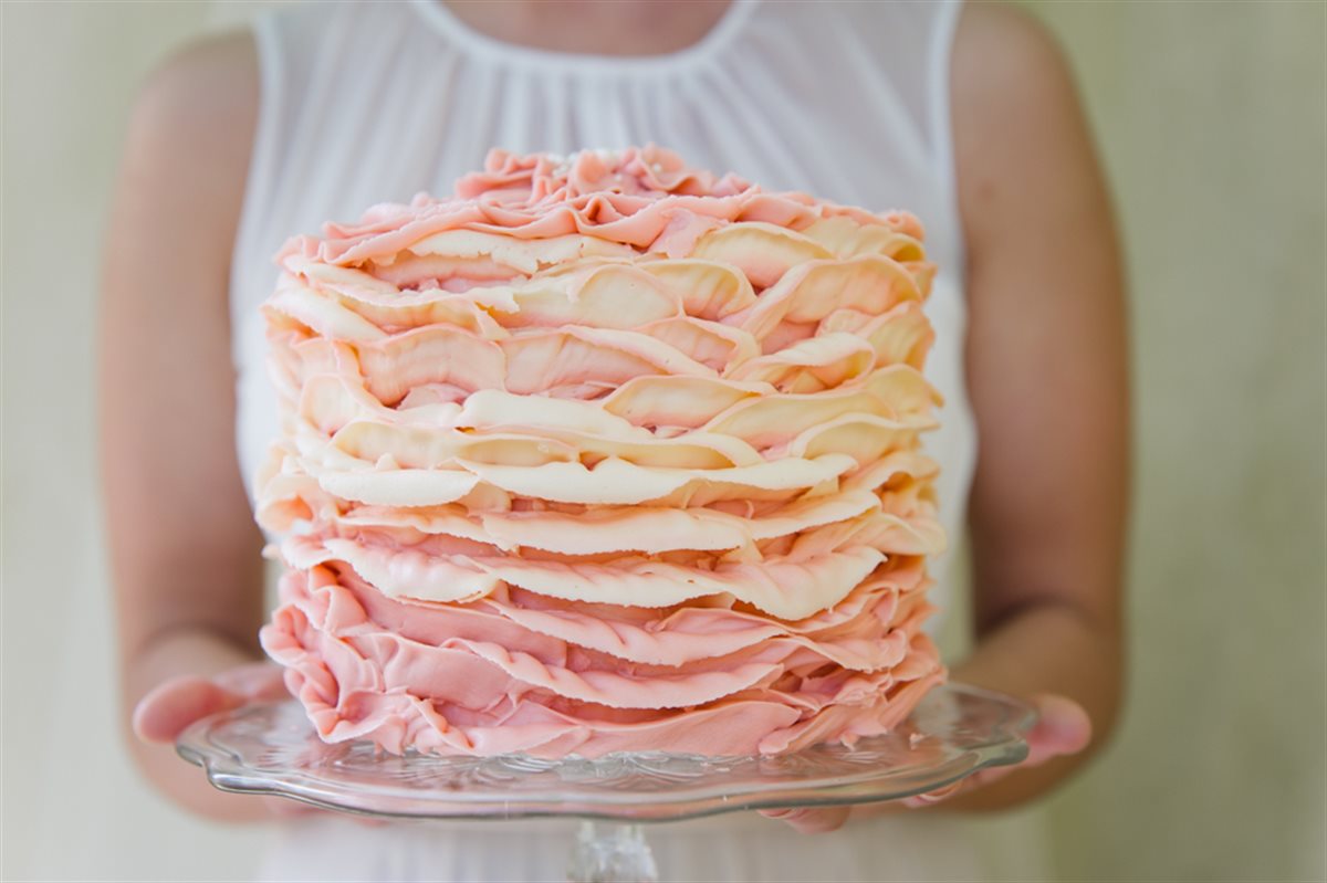 Editorial shot of a single-tier soft pink and cream buttercream ruffle celebration cake held in two hands on a glass pedestal, in soft natural light