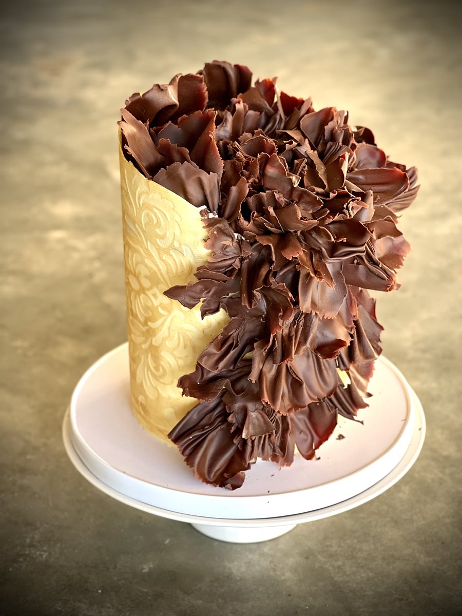 Tall single-tier celebration cake with a cream damask-patterned base half-covered in dramatic dark couverture chocolate ruffles cascading down one side