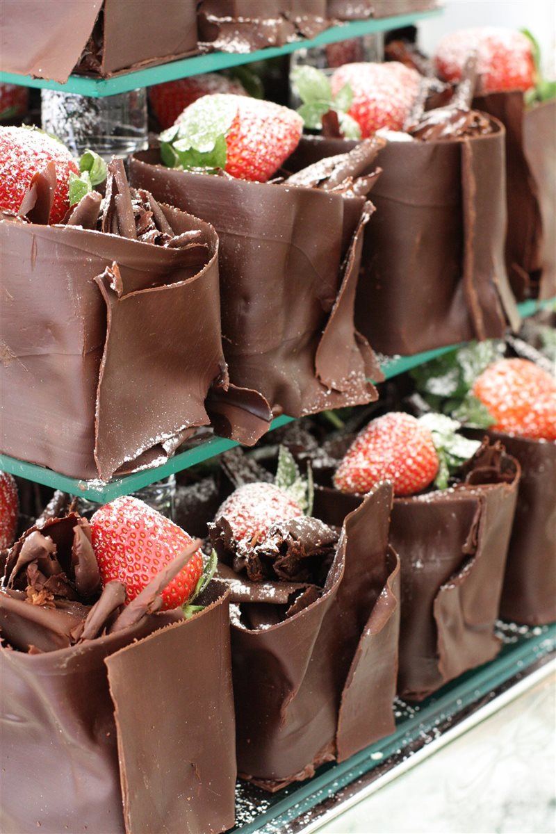 Individual dark couverture chocolate wrapped cakes filled with ganache, each nestled alongside a powdered-sugar strawberry on a display stand