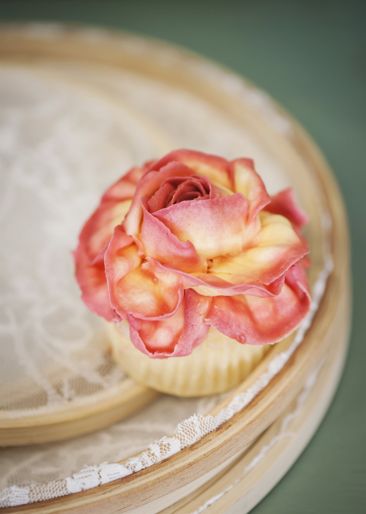 A single petite cupcake topped with a hand-piped peach and coral rose in full bloom, resting on a wooden tray with vintage lace, soft natural light against a deep green wall