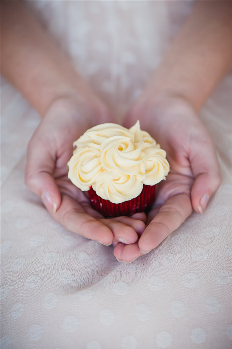 Hands gently holding a delicate red velvet cupcake decorated with cream cheese buttercream icing swirls — a petite gifting moment