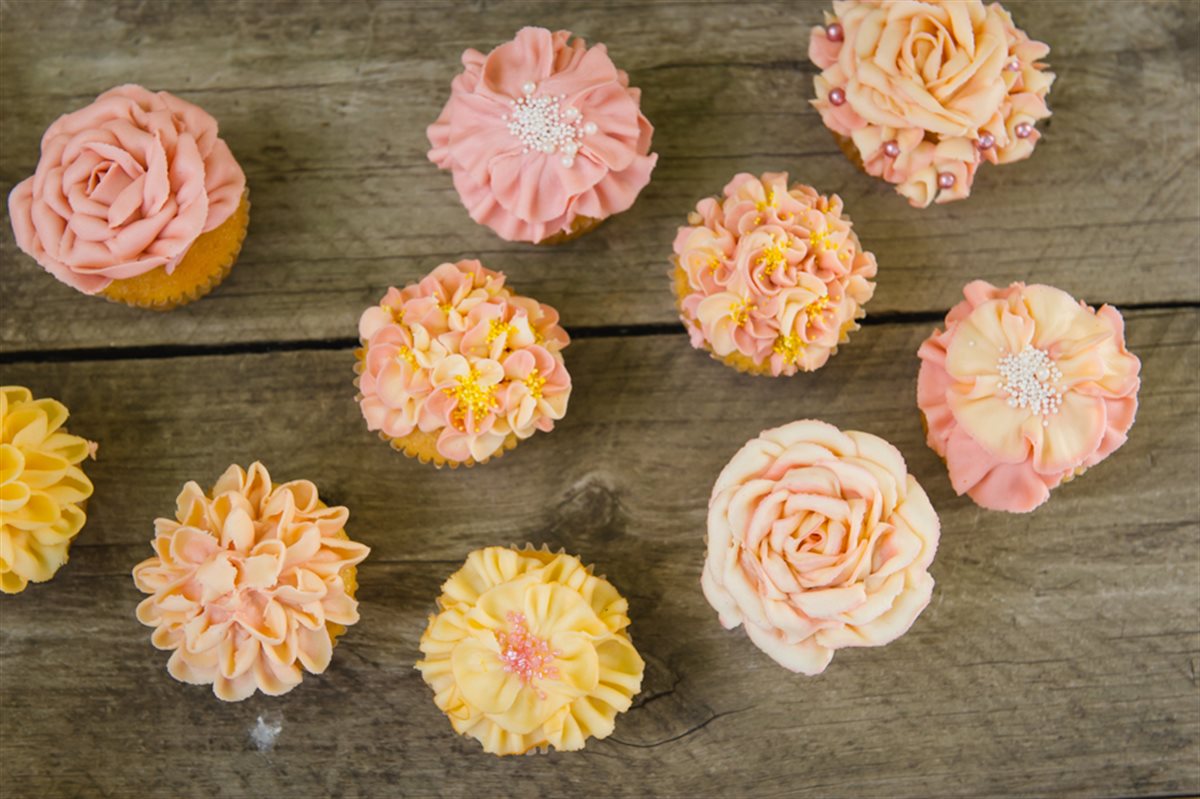 Overhead shot of nine buttercream cupcakes scattered across a rustic wooden table, in a soft peach, coral and yellow palette — hand-piped roses, dahlias and chrysanthemums