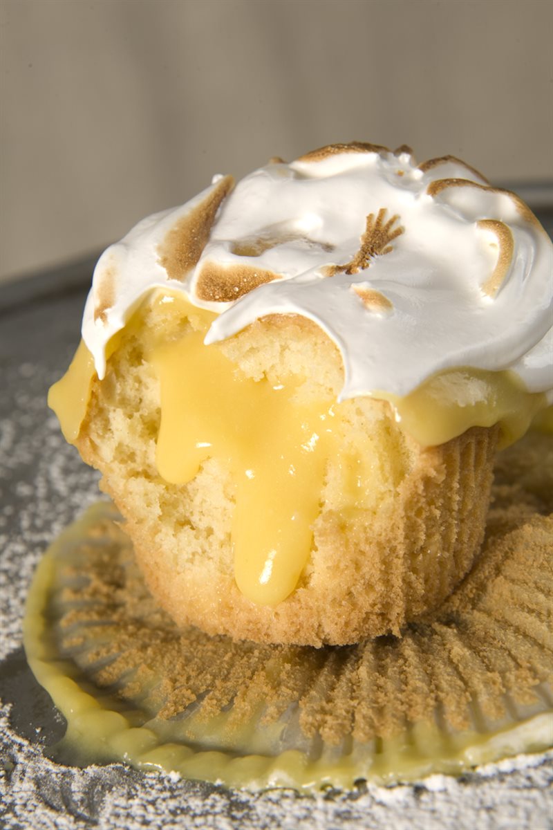 Petite lemon-meringue cupcake cracked open with lemon curd oozing from the centre and lightly torched Italian meringue on top, on a soft dusted surface