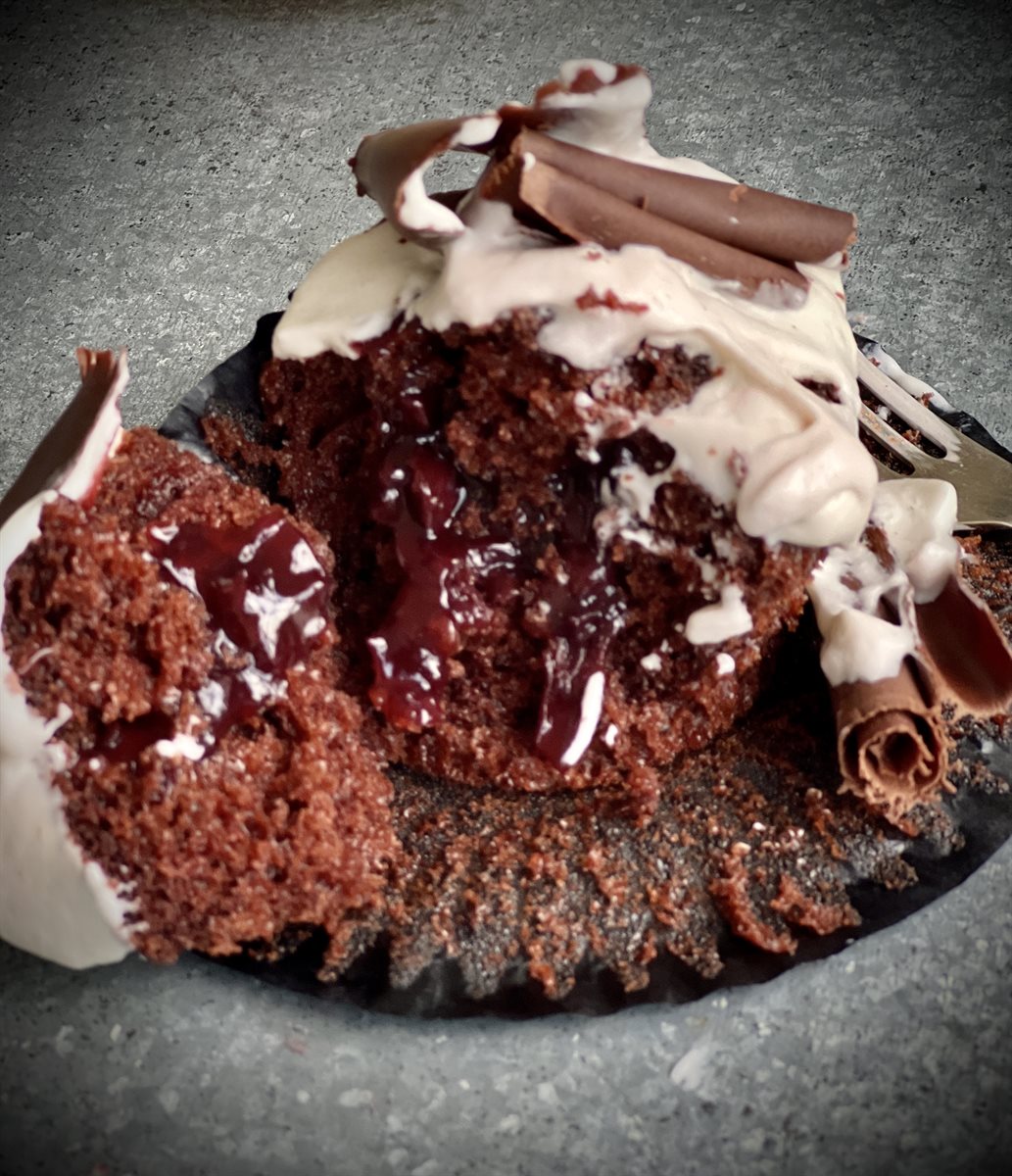 Dark chocolate cupcake cracked open with cherry compote spilling out, topped with cream and a couverture chocolate curl, on a concrete surface