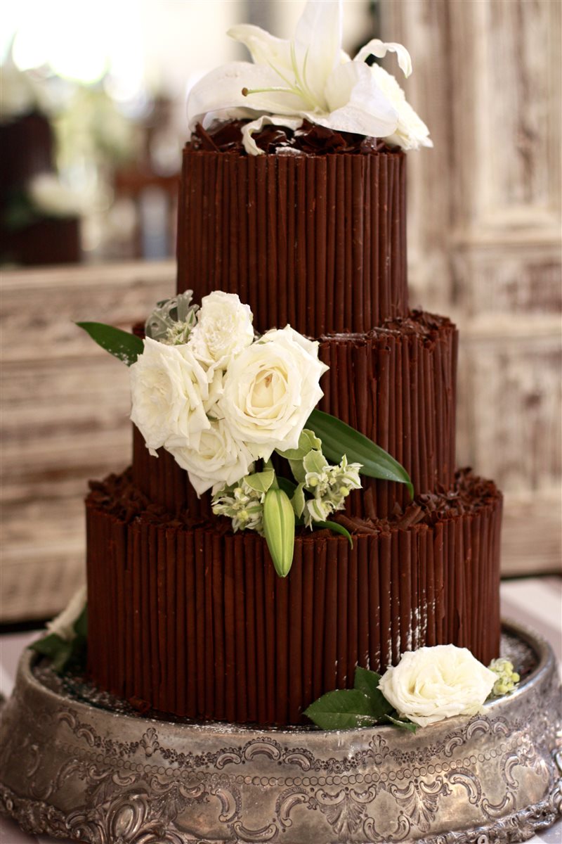 Three-tier dark chocolate cigar cake with clusters of white garden roses and a white lily on each tier, on an ornate silver cake stand in a softly lit venue