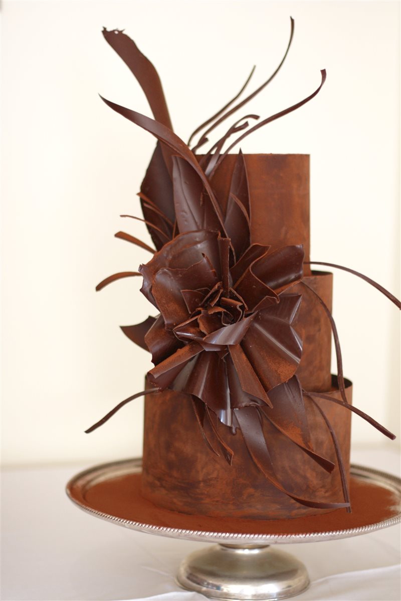 Three tiered smooth dark chocolate collar cake, dusted with cocoa, with a dramatic sculpted chocolate leaf-and-bloom explosion and wild chocolate ribbons rising from the surface, on a silver tray