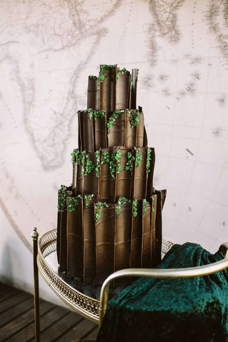 Four-tier dark chocolate vertical-scroll wedding cake with green sugar crystals, on brass tray with vintage Africa map backdrop