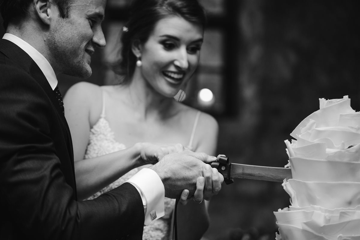 Bride laughing as she cuts her white couverture petal cake in a Cape barn — black & white