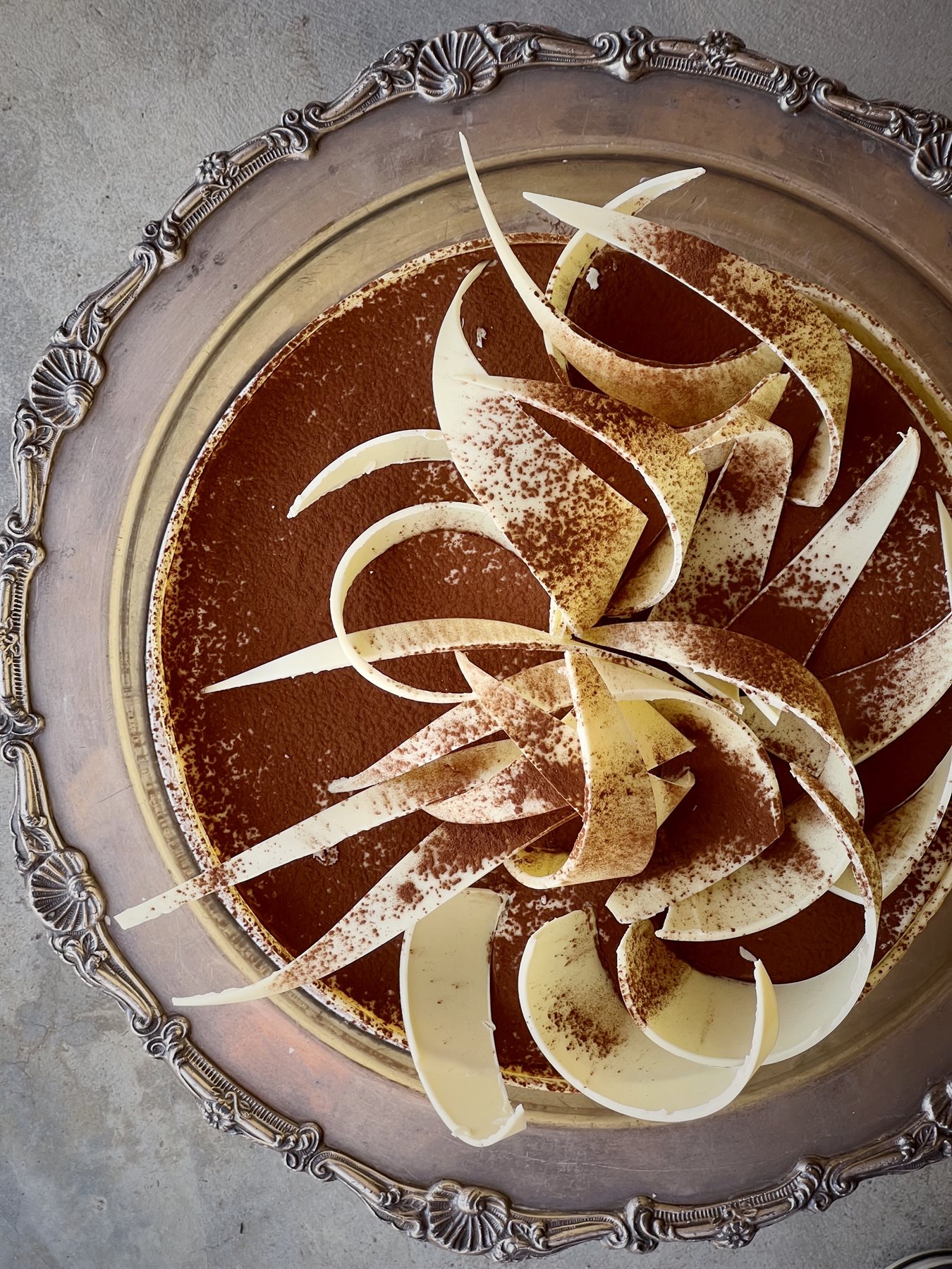 Aerial view of a single-tier chocolate cake with white chocolate curls fanned out in a spiral rosette, dusted with cocoa powder, on an ornate silver tray