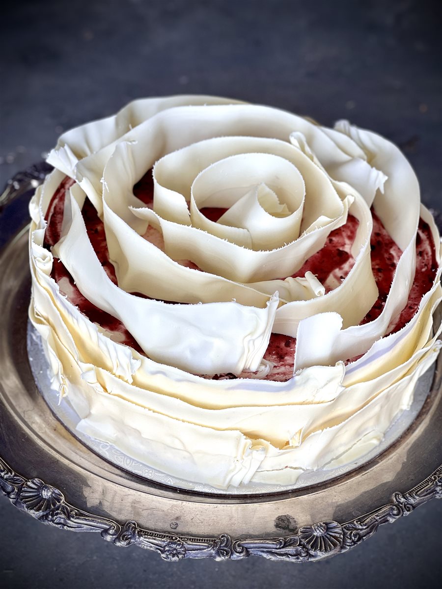 Single-tier cherry mascarpone cake wrapped and topped entirely in spiraling white couverture petals forming a full rose, with vivid red sponge visible through the white layers, on an ornate silver plate
