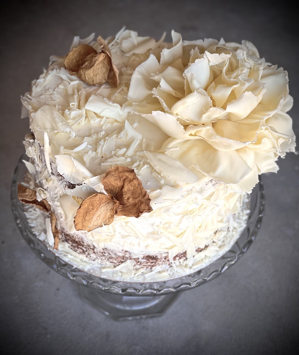 Single-tier semi-naked Hummingbird cake topped with a large sculpted white couverture chocolate bloom and an array of pale white petals, accented by dried autumn leaves, on a crystal glass cake stand