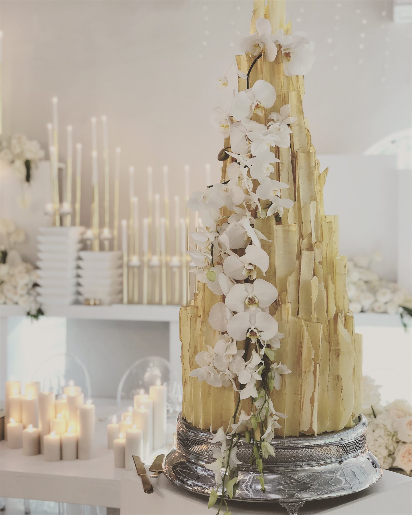 Multi-tier gold-touched white couverture wedding cake with cascading white orchids, on an ornate silver pedestal in a luxury candlelit venue setting