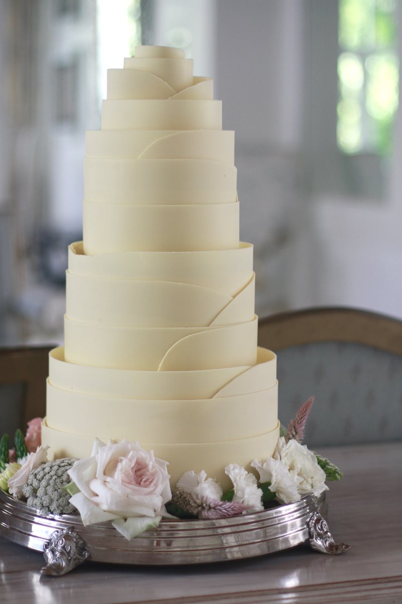 Four-tier white couverture spiral multiple white chocolate collar wedding cake with blush fresh roses and mixed flowers on silver stand, soft window light