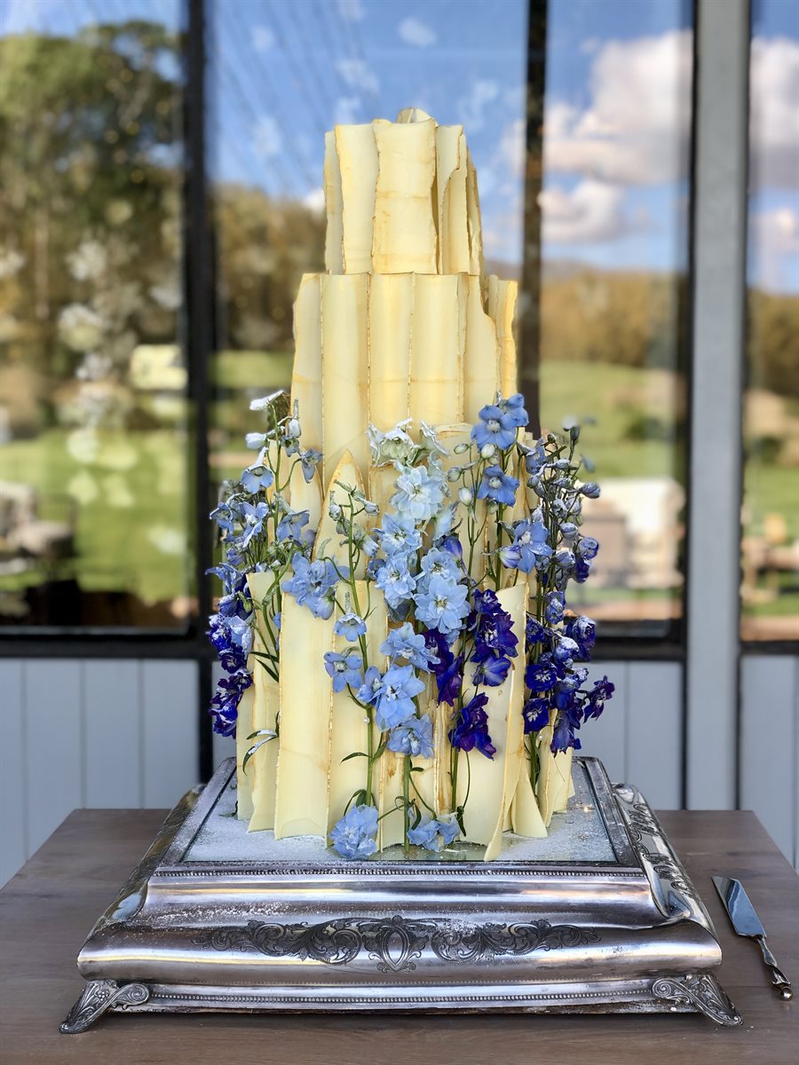 Tall architectural white chocolate shard cake, edges touched with gold and with cascading blue delphiniums on grand silver platform, garden venue beyond