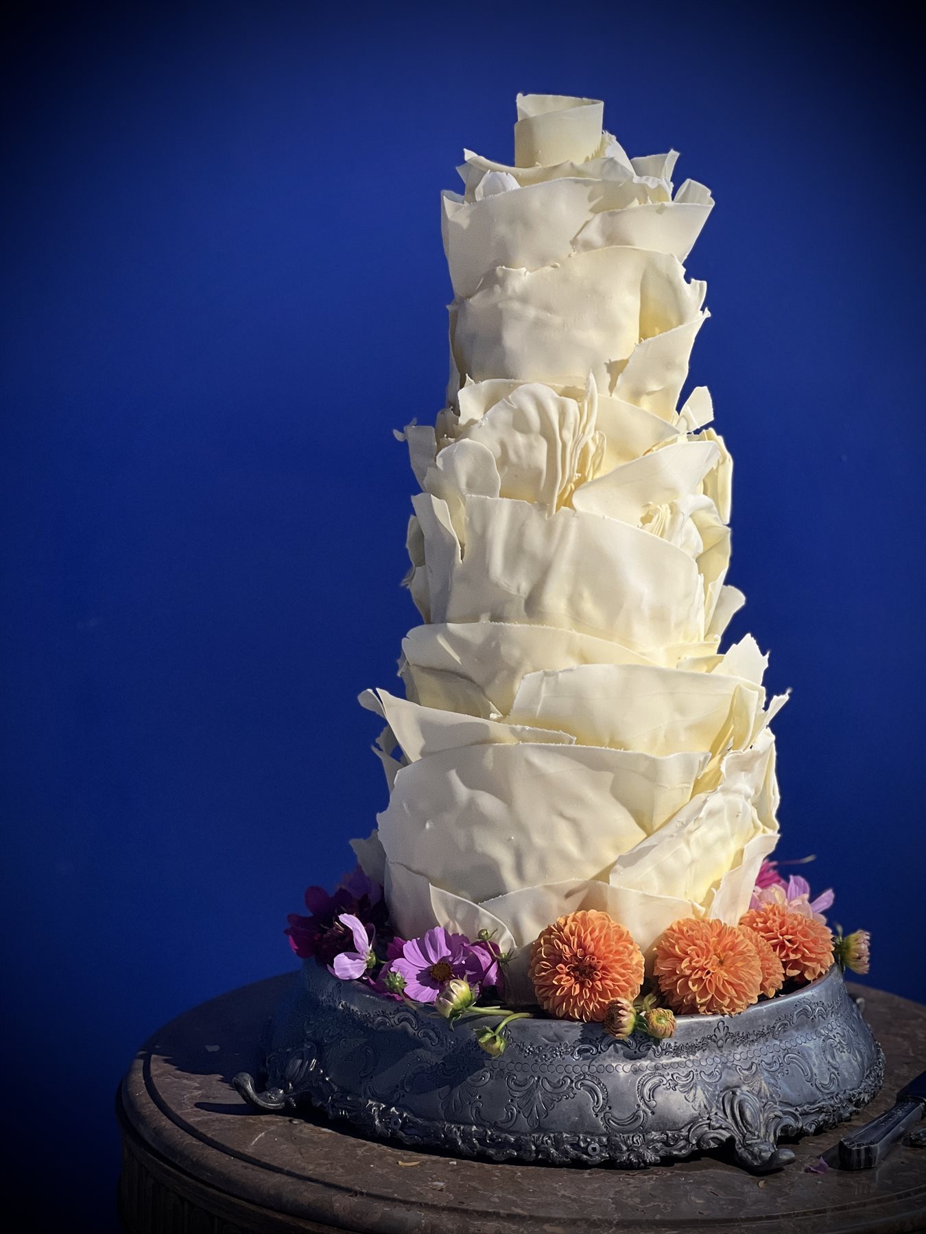 Five-tier pure white couverture petal cake against bold cobalt blue with orange dahlias