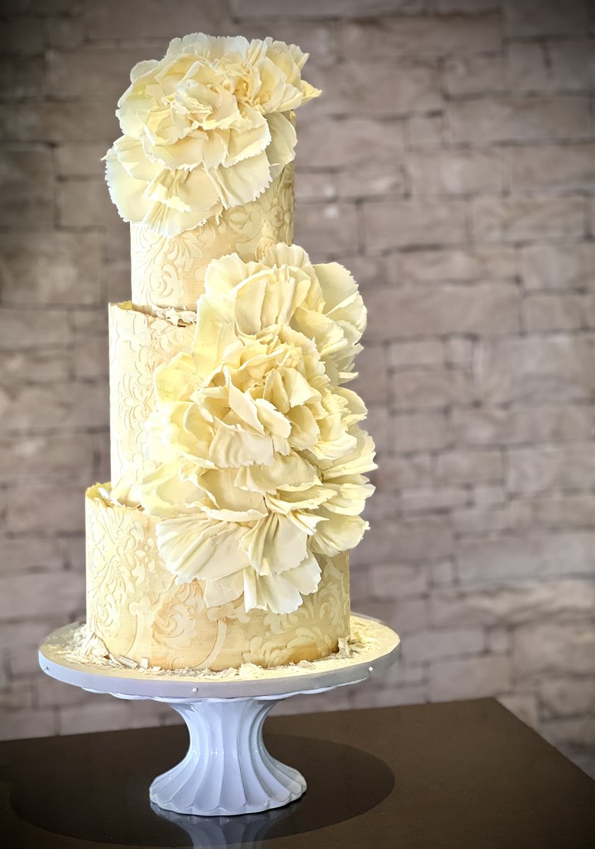 Three-tier white couverture cake with embossed lace damask pattern, lightly enhanced with gold paint, and large sculpted peony flowers cascading down, brick wall