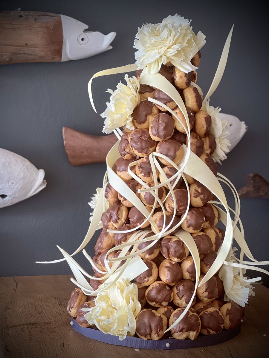 Tall croquembouche tower of chocolate-dipped profiteroles, draped with cascading white couverture chocolate ribbons and crowned with white chocolate carnation flowers, on a deep charcoal-grey background